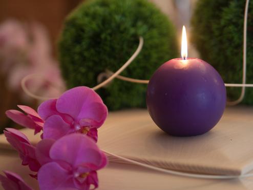 Lila Kerze mit Flamme, umgeben von pinken Orchideenblüten und dekorativen Pflanzen auf einem Tisch.