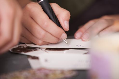 Nahaufnahme einer Hand, die mit einem Kugelschreiber auf einem Zettel schreibt, im Hintergrund eine brennende Kerze.
