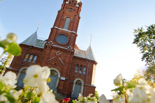 Nahaufnahme einer roten Backsteinkirche mit hohem Turm, umgeben von blühenden weißen Blumen im Vordergrund.