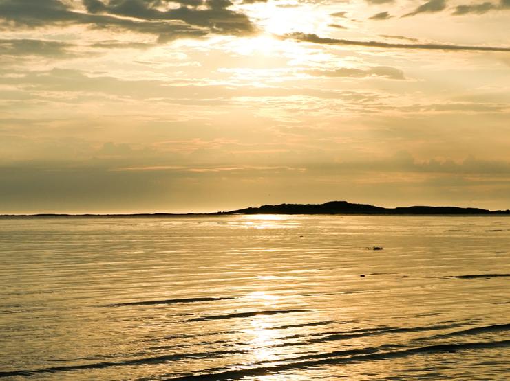 Sonnenuntergang über ruhiger Wasseroberfläche mit sanften Wellen und reflektierendem Licht. Im Hintergrund eine kleine Insel.